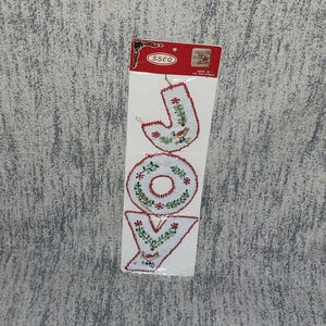 May include: A package of Christmas ornaments spelling out the word "JOY". The ornaments are white with red and green embroidered floral and bird designs. The package has a red header with the brand name "SSCO".