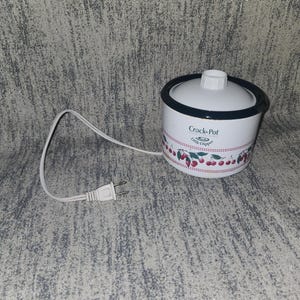 May include: A white Crock-Pot Little Dipper slow cooker with a black rim and a white lid featuring a knob. The exterior displays a red cherry pattern with green leaves and a red dotted line. A white electrical cord with a two-prong plug is attached.