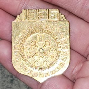 May include: A gold-colored metal medallion with the year "1986" at the top. The medallion features a circular design with letters and symbols, and a shield-like shape at the bottom. The medallion is held in a hand.