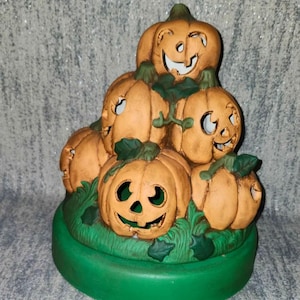May include: A ceramic Halloween decoration featuring a stack of orange pumpkins with carved faces. The pumpkins are arranged on a green base with green leaves. The pumpkins have various expressions.