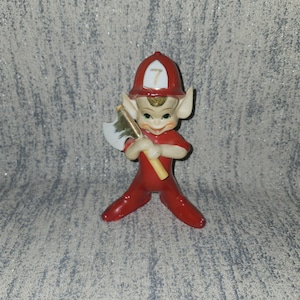 May include: A ceramic figurine of an elf in a red firefighter uniform, including a helmet with the number 7. The elf is holding a small axe with a green and white design. The figurine has large ears and a smiling expression.