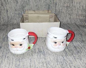 May include: Two ceramic Santa Claus mugs with white faces, red handles, and holly accents. The mugs are white with a red handle and a Santa Claus face design. The mugs are next to a cardboard box.