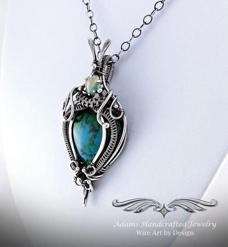 May include: A silver wire wrapped pendant featuring a green and blue gemstone with a smaller white opal gemstone above it. The pendant is attached to a silver chain.