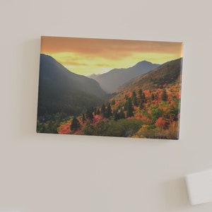 Könnte beinhalten: Ein Foto einer Bergkette mit einem Tal im Vordergrund. Die Berge sind mit Bäumen bedeckt, und das Tal ist mit Bäumen gefüllt, die ihre Farbe wechseln. Der Himmel ist leuchtend orange und gelb, und die Sonne geht hinter den Bergen unter.