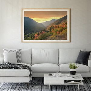 May include: A framed photograph of a mountain valley with fall foliage in shades of red, orange, and yellow. The sky is a soft orange and yellow, and the mountains are covered in trees.