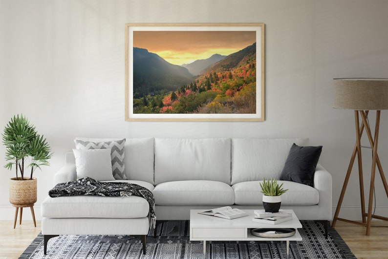 May include: A framed photograph of a mountain valley with fall foliage in shades of red, orange, and yellow. The sky is a soft orange and yellow, and the mountains are covered in trees.