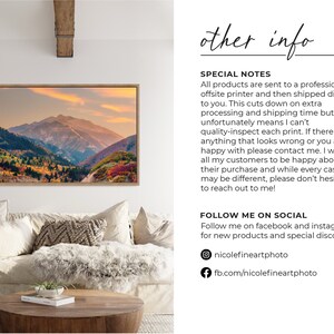 May include: A framed photograph of a mountain range with fall foliage in shades of red, orange, and yellow. The mountains are in the background, and the foreground is a valley with trees and a road. The sky is a soft blue with a few clouds. The photograph is hanging on a white wall in a living room setting.