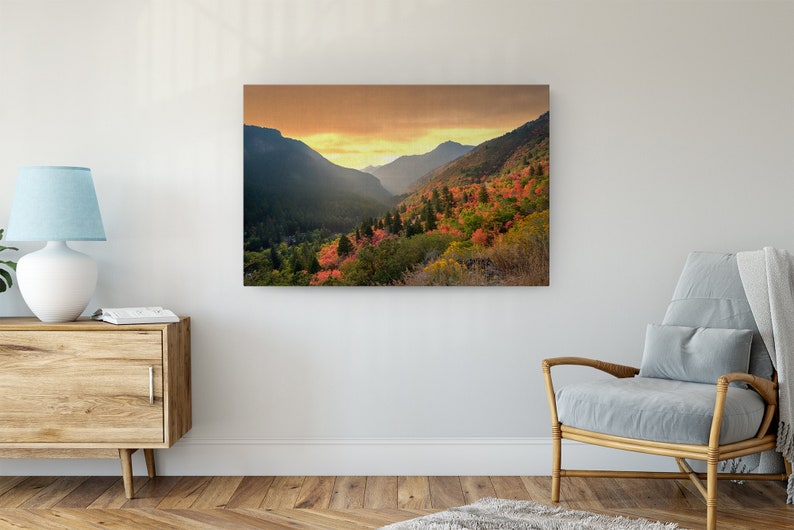 May include: A scenic landscape photograph of a mountain valley with a golden sunset in the background. The valley is filled with trees, many of which are showing fall colors of red, orange, and yellow.