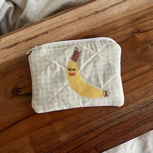 May include: A small, quilted coin purse with a light blue and yellow checkered pattern. The purse features a smiling banana applique with brown and yellow details. The zipper is white.