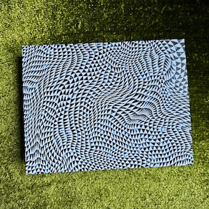 May include: A rectangular item with a black and white geometric pattern of triangles, with hints of light blue. The item is resting on a green, textured surface. The pattern creates a wavy, three-dimensional effect.
