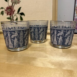 May include: Three clear glass tumblers with a navy blue band featuring a white classical design. The glasses are arranged on a light brown wooden surface. The design includes figures and architectural elements.