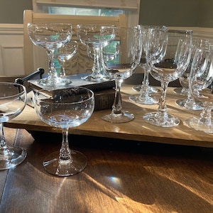 May include: A set of nine clear glass stemmed drinking glasses, including champagne flutes, wine glasses, and coupe glasses, arranged on a wooden tray.