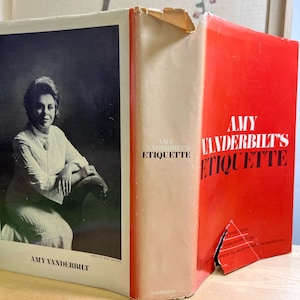 May include: A red hardcover book titled "Amy Vanderbilt's Etiquette" with a black and white photo of Amy Vanderbilt on the front cover. The book is slightly damaged on the spine.