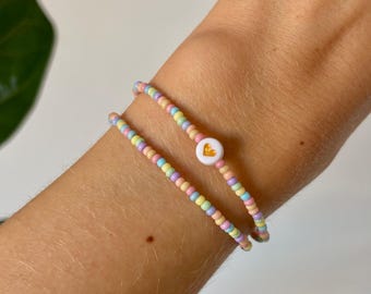May include: Two pastel-colored beaded bracelets with a white heart charm. The bracelets are layered on a person's wrist.