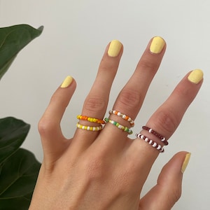 Könnte beinhalten: Eine Hand zeigt vier Perlenringe in Orange, Gelb, Grün und Braun. Jeder Ring besteht aus kleinen, farbigen Perlen. Die Ringe werden an verschiedenen Fingern getragen und zeigen die Vielfalt der Designs und Farben.
