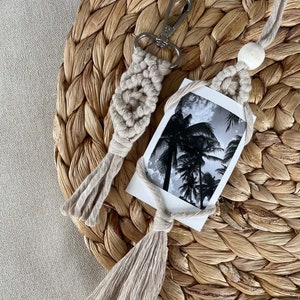 May include: A beige macrame keychain with a silver clasp and a white wooden bead. The keychain is attached to a white card with a black and white photo of palm trees.