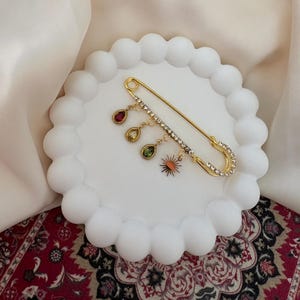 Könnte beinhalten: Eine goldfarbene Sicherheitsnadel-Brosche mit klaren Strasssteinen und baumelnden Anhängern. Die Anhänger umfassen einen roten Tropfen, einen grünen Tropfen und einen kleinen Sonnenaufgang. Die Brosche wird auf einem weißen Teller mit gewelltem Rand präsentiert.