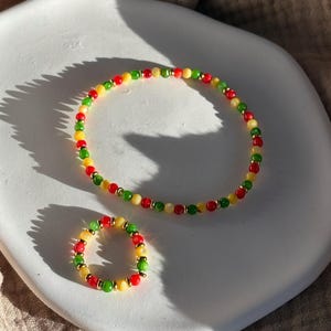 Może przedstawiać: Zestaw bransoletki i pierścionka z koralików. Biżuteria zawiera czerwone, żółte, zielone i złote koraliki. Bransoletka ma okrągły kształt, a pierścionek jest mniejszą wersją bransoletki. Biżuteria jest prezentowana na białym, okrągłym talerzu.