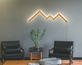 May include: A living room interior with two black leather armchairs, a wooden coffee table, and a wooden floor. A potted plant sits on a small side table. A geometric light fixture in the shape of a mountain range is mounted on the wall.