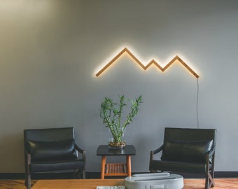 May include: A living room interior with two black leather chairs, a wooden coffee table, and a wooden floor. A potted plant sits on a small side table. A geometric light fixture in the shape of a mountain range is mounted on the wall.