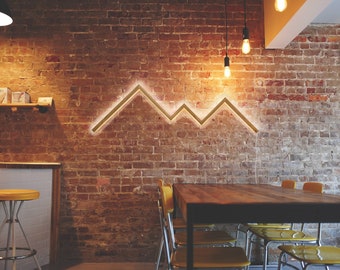 May include: A wooden table with yellow chairs sits in a restaurant with a brick wall. A gold metal mountain-shaped light fixture is mounted on the wall.