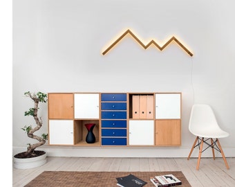 May include: A white wall with a wooden geometric light fixture shaped like a mountain range. Below the light fixture is a white and wood cabinet with blue drawers. A white chair with wooden legs is on the right side of the image.