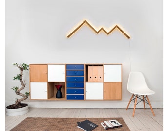 May include: A white wall with a wooden geometric light fixture shaped like mountains. Below the light fixture is a white and wood cabinet with blue drawers. A white chair with wooden legs is in the corner of the room.