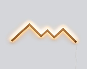 May include: A wooden wall light shaped like a mountain range with a warm white glow. The light is wired with a cord.