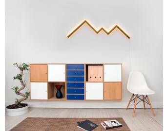 May include: A white chair with wooden legs sits in front of a modular cabinet with white, blue, and wood doors. A wooden light fixture shaped like a mountain range is mounted on the wall behind the cabinet.