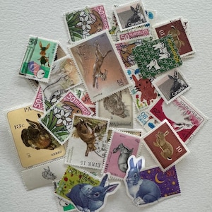 Könnte beinhalten: Eine Sammlung alter Briefmarken mit verschiedenen Hasenillustrationen. Die Briefmarken zeigen unterschiedliche Farben, Designs und Herkunftsländer, darunter Briefmarken aus Kanada, Japan und Österreich. Die Briefmarken sind auf einer weißen Oberfläche angeordnet.