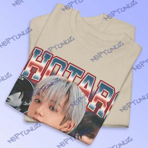 Puede incluir: Camiseta beige con un gr&aacute;fico que presenta la palabra "HOTARU" en letras rojas, blancas y azules, junto con un primer plano del rostro de una persona. La camiseta est&aacute; doblada, con el gr&aacute;fico visible en la capa superior.