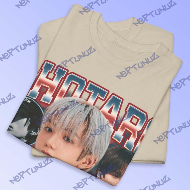 Puede incluir: Camiseta beige con un gr&aacute;fico que presenta la palabra "HOTARU" en letras rojas, blancas y azules, junto con un primer plano del rostro de una persona. La camiseta est&aacute; doblada, con el gr&aacute;fico visible en la capa superior.