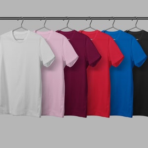 May include: A row of six solid-color t-shirts hanging on a metal rod. The shirts are white, light pink, burgundy, red, blue, and black. Each shirt has short sleeves and a crew neck.