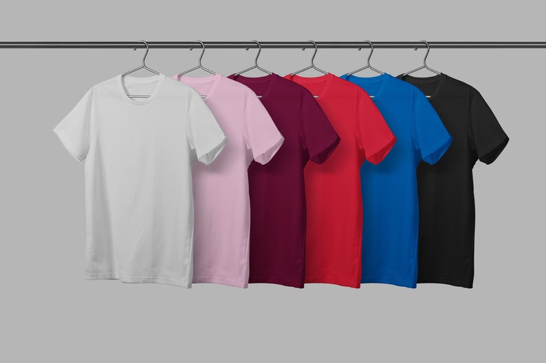 May include: A row of six solid-color t-shirts hanging on a metal rod. The shirts are white, light pink, burgundy, red, blue, and black. Each shirt has short sleeves and a crew neck.