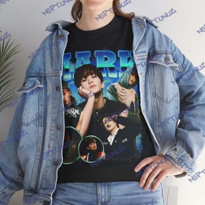 Op de afbeelding: Zwart T-shirt met een grafische print van een groep mannen. De grafiek is in retro-stijl met een blauw en groen kleurenschema. De tekst "MARK" is in grote letters bovenaan de grafiek gedrukt.
