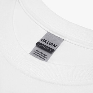 May include: Close-up of a white t-shirt collar with a gray tag. The tag reads "GILDAN HEAVY COTTON" and includes text indicating the shirt was made in Nicaragua in English, French, and Spanish.