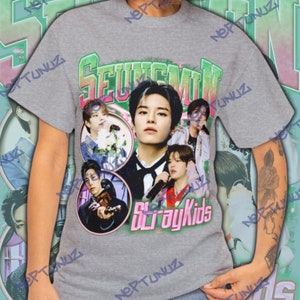 Puede incluir: Una camiseta gris con un collage de im&aacute;genes de un artista musical, con el nombre "SEUNGMIN" en letras verdes y rosas. Las palabras "Stray Kids" est&aacute;n impresas en rosa debajo de las im&aacute;genes. La camiseta tiene un dise&ntilde;o vintage y retro.