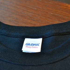 Puede incluir: Primer plano de un cuello de camiseta negra con una etiqueta blanca. La etiqueta dice "GILDAN Heavy Cotton" en texto azul y rojo. La camiseta est&aacute; hecha de una tela oscura y suave.