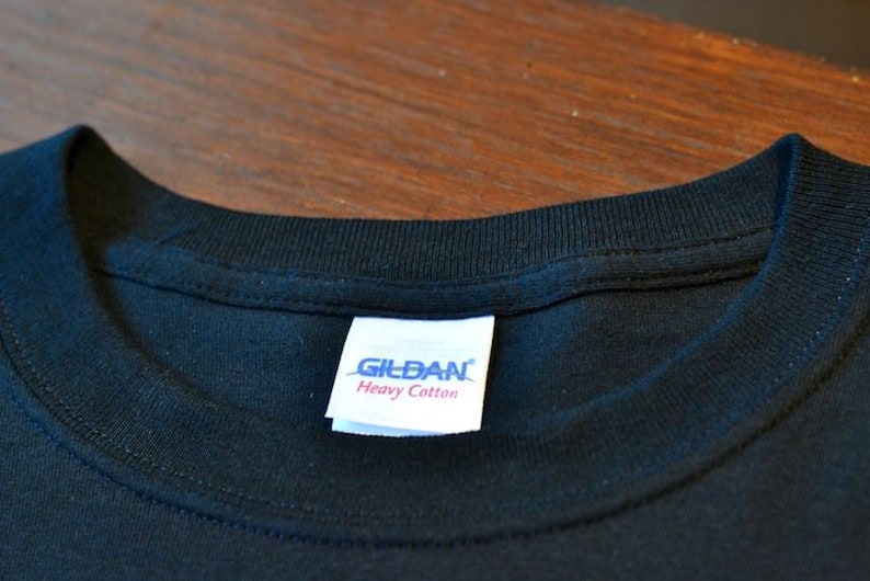 Puede incluir: Primer plano de un cuello de camiseta negra con una etiqueta blanca. La etiqueta dice "GILDAN Heavy Cotton" en texto azul y rojo. La camiseta est&aacute; hecha de una tela oscura y suave.