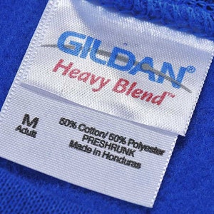 K&ouml;nnte beinhalten: Nahaufnahme eines wei&szlig;en Etiketts mit dem Markennamen "GILDAN" in Blau und "Heavy Blend" in Rot. Das Etikett gibt auch die Gr&ouml;&szlig;e "M Adult" und die Stoffzusammensetzung an: 50% Baumwolle und 50% Polyester. Das Etikett ist auf einem blauen Stoff befestigt.