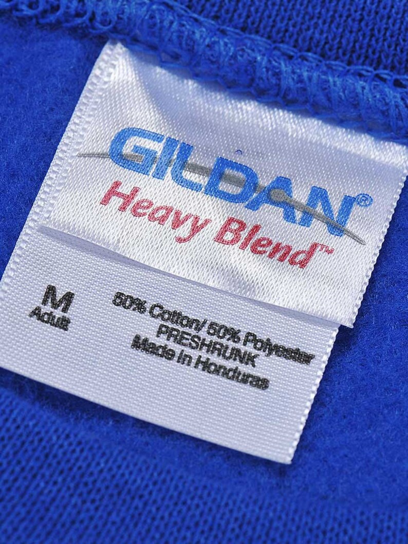 K&ouml;nnte beinhalten: Nahaufnahme eines wei&szlig;en Etiketts mit dem Markennamen "GILDAN" in Blau und "Heavy Blend" in Rot. Das Etikett gibt auch die Gr&ouml;&szlig;e "M Adult" und die Stoffzusammensetzung an: 50% Baumwolle und 50% Polyester. Das Etikett ist auf einem blauen Stoff befestigt.