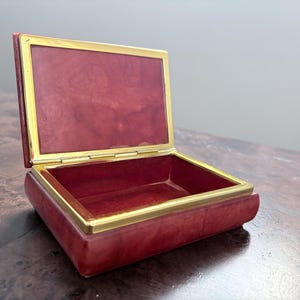 May include: Open red jewelry box with a gold-colored trim. The box is rectangular with a hinged lid. The interior is lined with a matching red material.