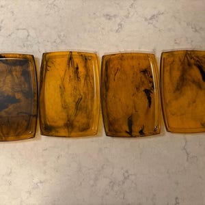 May include: Four rectangular coasters with rounded corners. The coasters have a translucent amber color with dark brown marbling patterns. The coasters are arranged in a row, showcasing their unique design and color variations.
