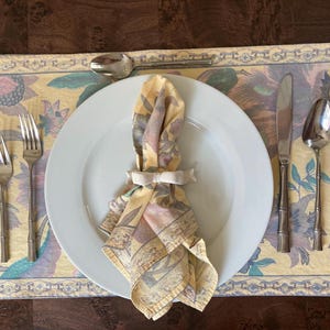 May include: A white dinner plate with a folded floral napkin, two forks, a spoon, and a knife on a yellow floral placemat.