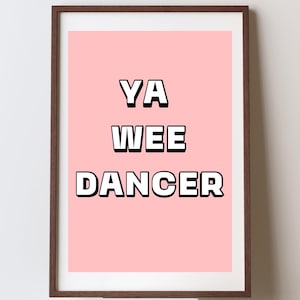 May include: A framed art print with the text "YA WEE DANCER" in bold white letters with black outlines, set against a light pink background. The frame is a dark brown color. The artwork is displayed on a white surface.