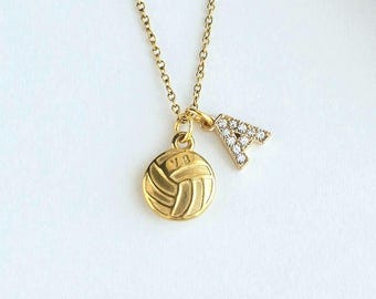 May include: A gold-toned necklace featuring a delicate chain with two charms: a circular gold volleyball pendant with embossed lines and a small, capital letter A charm encrusted with clear, sparkling stones. The jewelry piece is displayed clearly.