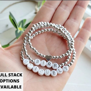 May include: A silver beaded bracelet with a heart charm and white beads spelling out the word "uniquely".