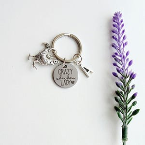 May include: A silver keychain with a chicken charm, a round tag with "CRAZY chicken LADY" text, and an "A" charm. A purple lavender stem is on the right side of the image. The keychain is on a white background.