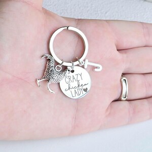 May include: Silver-toned keychain featuring a rooster charm and a round tag. The tag reads "Crazy Chicken Lady" with a heart symbol. The keychain is held in a hand.