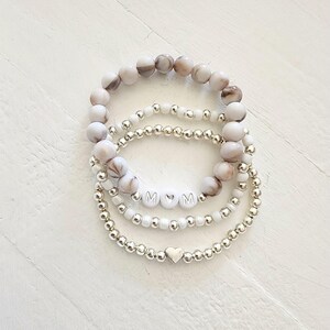 May include: Set of four bracelets with white and brown marbled beads, silver beads, and silver letter beads spelling "MOM" with a heart between the letters. The bottom bracelet has a silver heart charm.